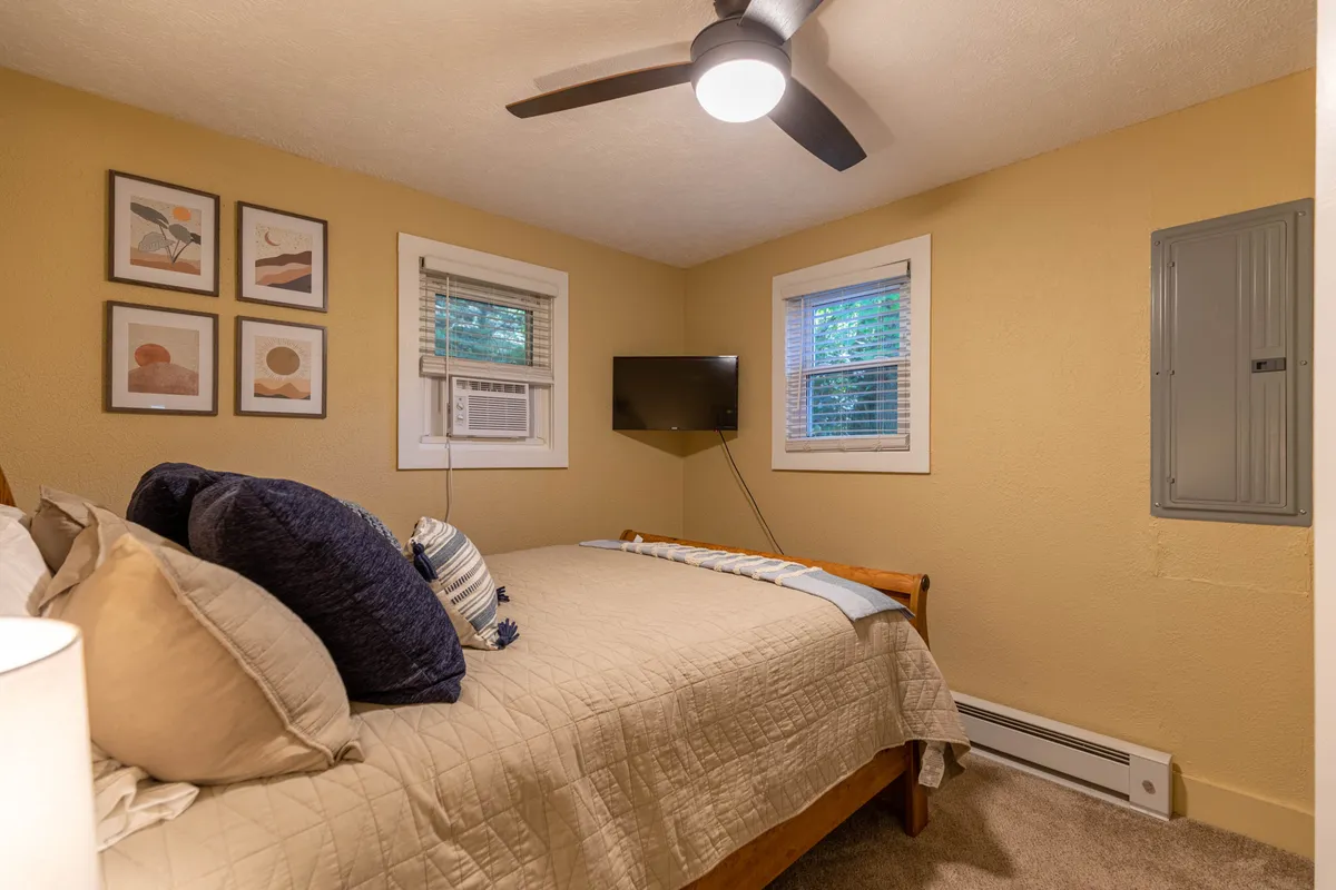 A welcoming bedroom with a cozy bed, decorative wall art, natural light pouring through windows, and a TV mounted on the wall. The ambiance is inviting, perfect for relaxing or enjoying a quiet night in.