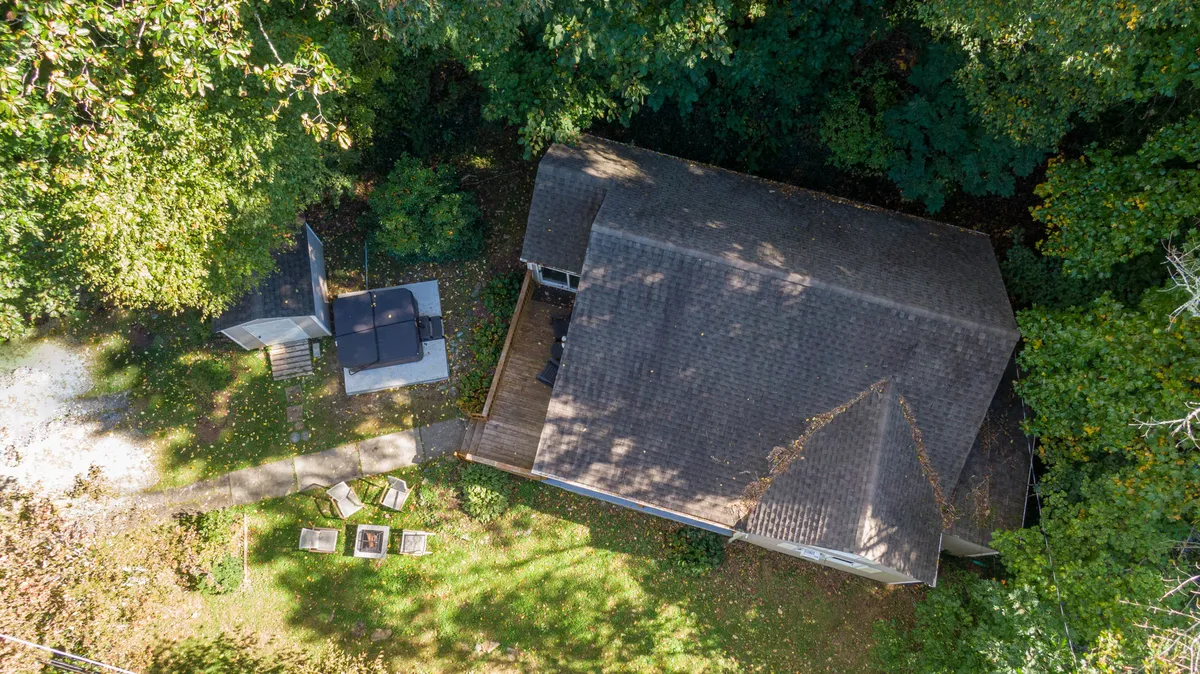 A view of the expansive deck area with patio furniture, a roof structure for shade, garden beds, and lush trees all around. The space feels tranquil and is clearly designed for gatherings and outdoor enjoyment.