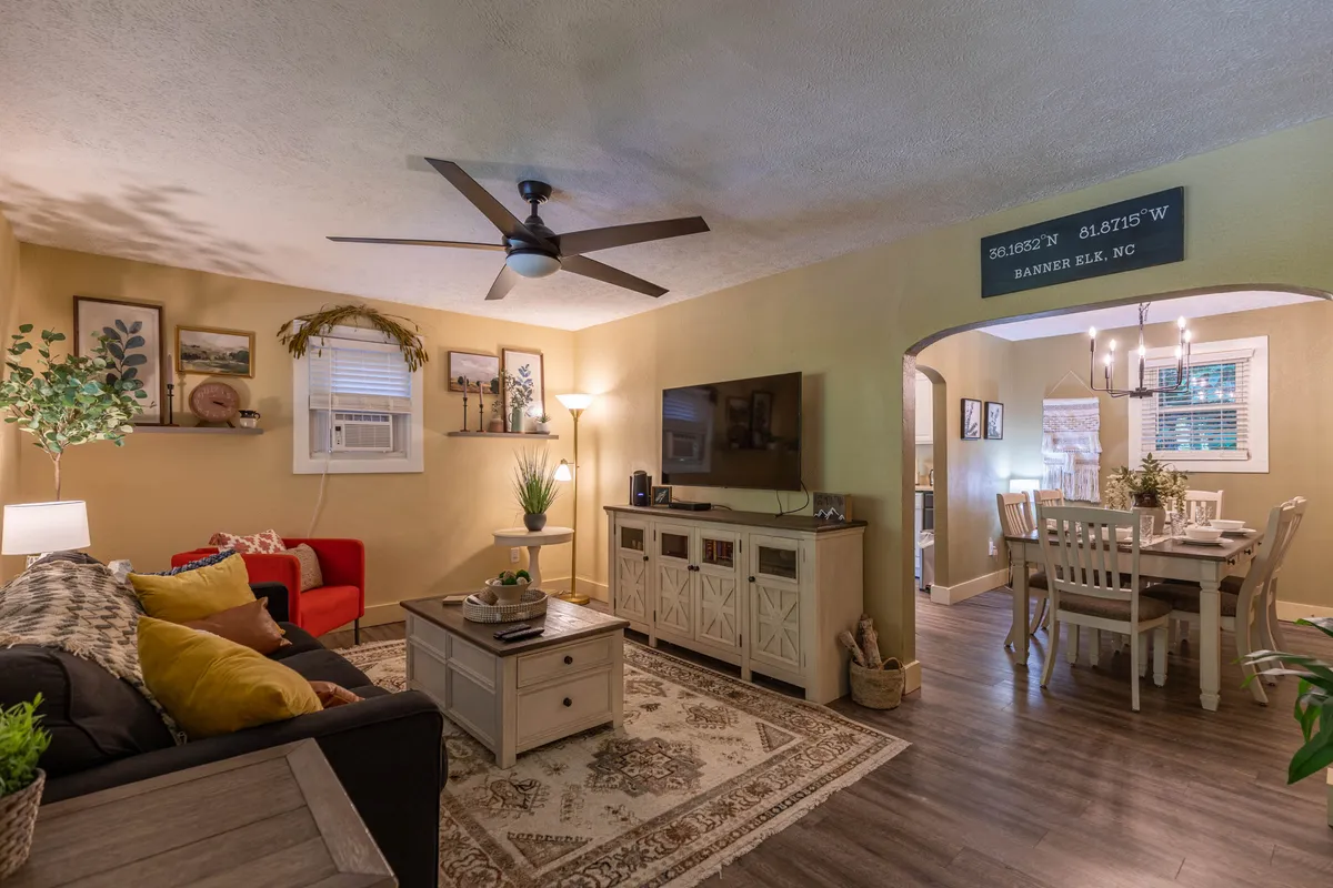 This image features a cozy living room with a sofa adorned with colorful cushions, a coffee table with decorative items, a modern TV unit, and the dining area visible in the background. The open layout hints at easy flow between lounging and gathering spaces.