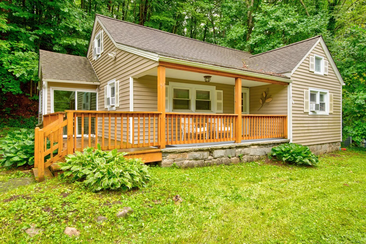 The image showcases the inviting exterior of Hot Toddy Cottage, featuring a charming wooden porch surrounded by green landscaping and a sturdy stone foundation. Dormer windows and cozy architectural details suggest a warm family retreat, with open space for children and pets to play.