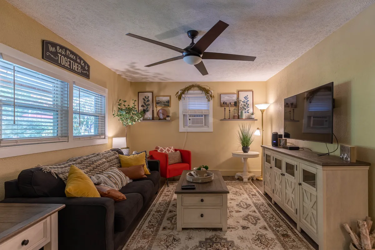 A plush living room scene with a sofa featuring colorful pillows, a red accent chair, decorative wall art, and a television unit. The cozy setup invites relaxation and socializing, with ample lighting and thoughtful decor.