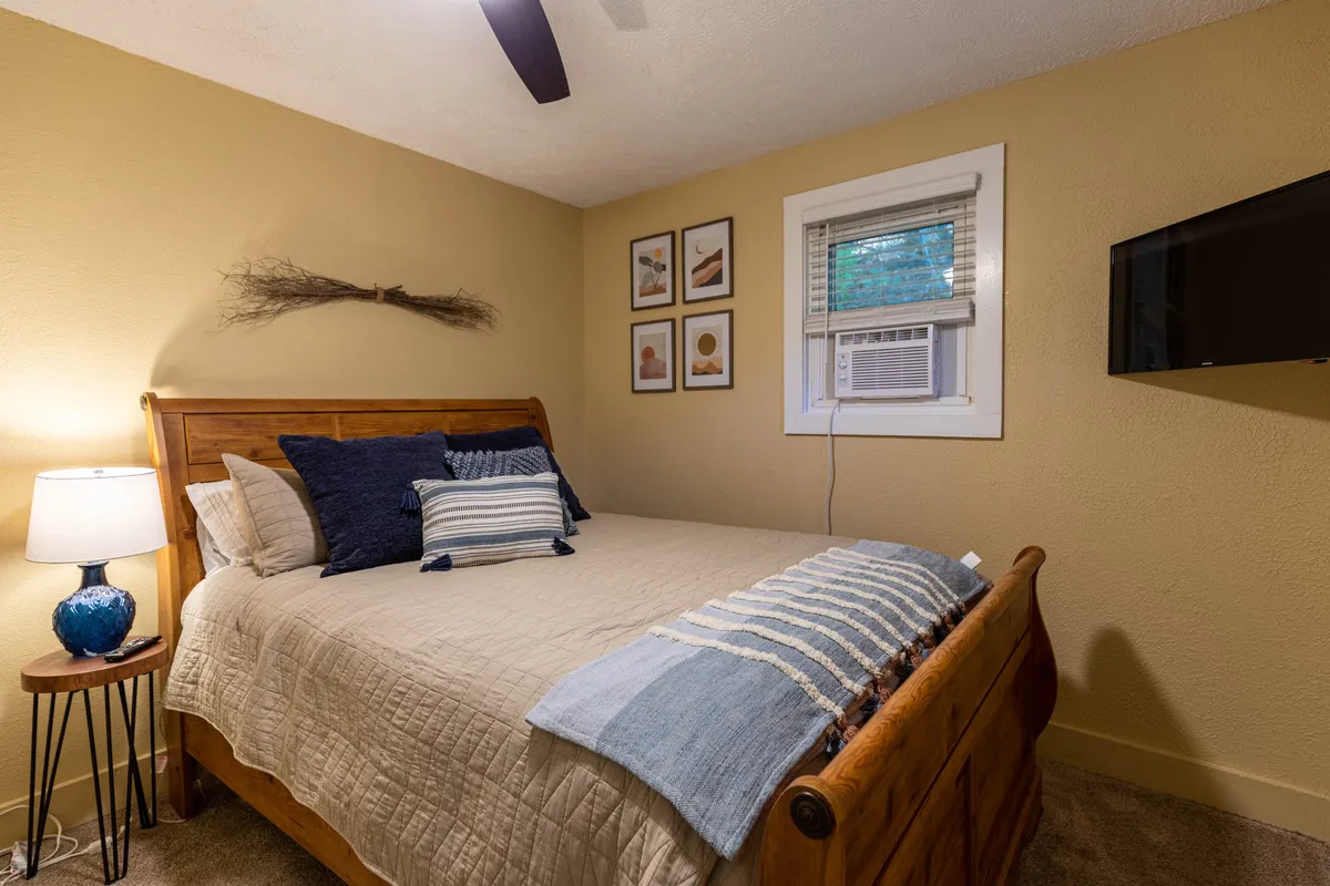 This image features a serene bedroom with a comfortable bed, decorative wall art, a bedside lamp, and a visible air conditioning unit. The atmosphere is tranquil and restful, emphasizing comfort and privacy.
