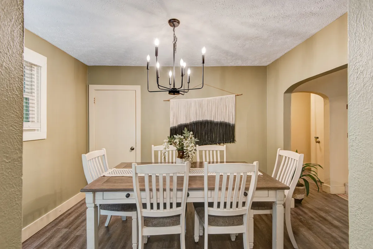 The dining table is set beneath a chandelier, surrounded by plants and a wall hanging, creating a warm and intimate space ideal for meals with friends or romantic dinners for two.