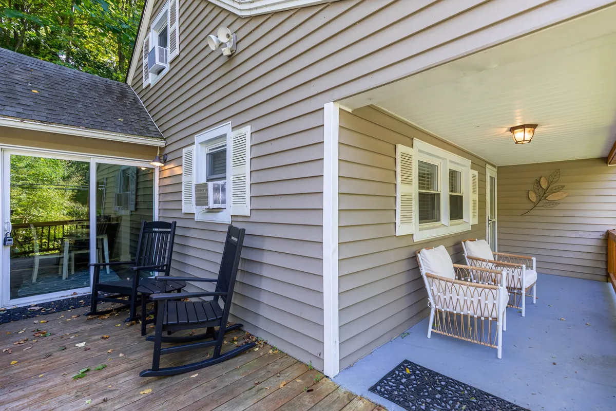 A front or side porch scene with cozy rocking and woven chairs, large windows, and lush natural surroundings. The image feels serene and inviting, suggesting spaces for quiet moments and enjoying the setting.